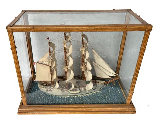 K1639 Late 19th century  early 20th century Ship Diorama in glass case depicting a three-masted ship with full sails flying an American Flag. The highly detailed, unnamed ship is made of what appears to be a salt glazed style pottery with stitched cloth sails and hand painted water. The glass panes (one has a corner crack) are enclosed in a removeable metal case that retains its mustard paint decorated surface. Case Measurements: 20 ¾ wide x 10 ¼ deep x 16 tall. 