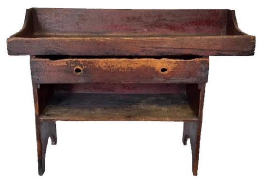 J217 Extremely rare Lancaster County, Pennsylvania bucket bench / dry sink featuring an overhanging well with a drawer and a mortised open shelf below the center section of the well. The surface retains traces of old gray paint over the original red paint, and bears extensive wear indicative of many years of repetitive use. The notched groove drain hole prominently remains in the overhang of the well. The drawer opens to reveal a divided section for utensils. One board, square head nail construction with nice, half-moon cut out ends.  This is one of the earliest, and best, examples of the Lancaster County bucket bench / dry sinks we have seen in 40+ years. Circa 1830�s � 1840�s. Measurements: 48 &frac34;� wide x 19 &frac12;� deep x 35 &frac14;� tall.